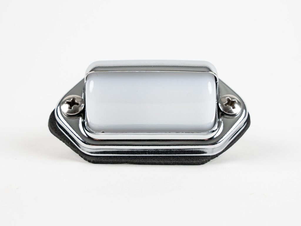 Front-angled view of a surface-mount chrome-hooded license plate light featuring a high-polish finish, two Phillips mounting screws, black foam mounting gasket, and wiring leads against a white background.