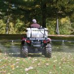 Workhorse 25-gallon sprayer mounted on the rear of a red ATV, actively spraying lawn with its extended 7-nozzle boom.